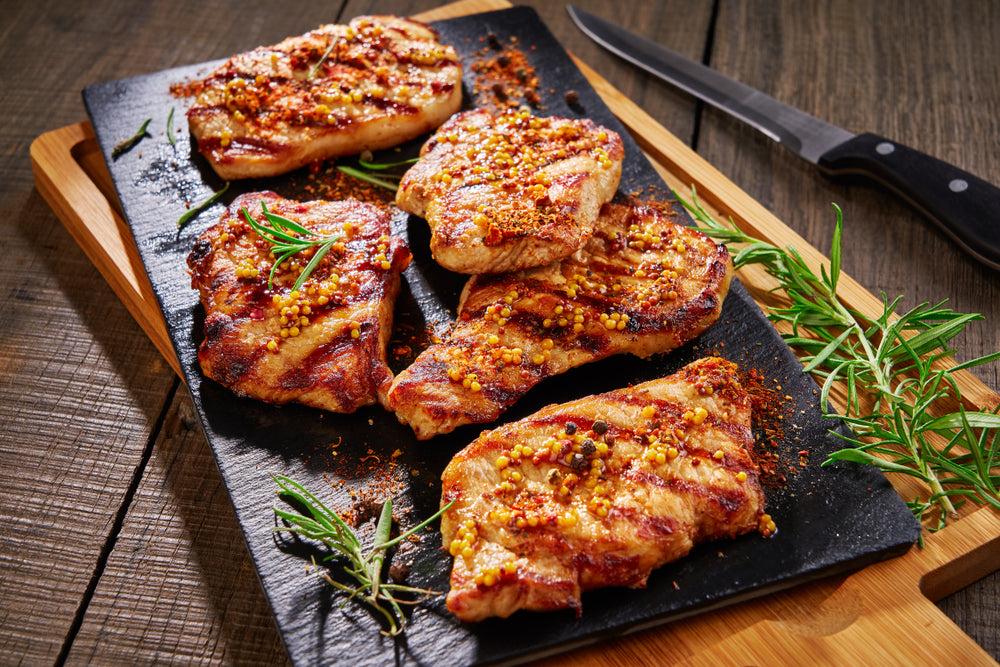Grilled pork chops seasoned with spices and mustard seeds are arranged on a black slate platter, garnished with fresh rosemary, on a wooden table alongside a knife.