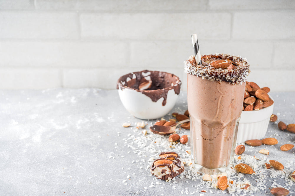 A tall glass of chocolate almond smoothie, topped with shredded coconut and an almond, stands next to a bowl of melted chocolate and scattered almonds on a gray countertop.