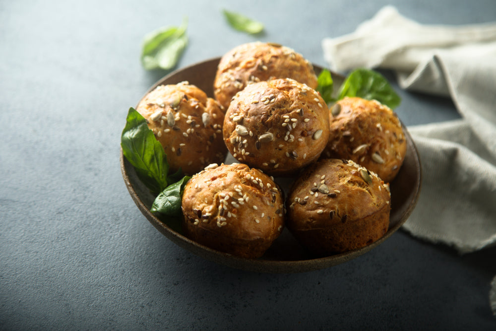 A bowl filled with six golden-brown pumpkin protein muffins topped with seeds sits on a textured gray surface, surrounded by fresh basil leaves and a light cloth.