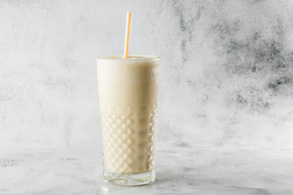 A tall glass of creamy peach pie shake with a yellow straw stands against a light gray background, showcasing its smooth texture and inviting appearance.