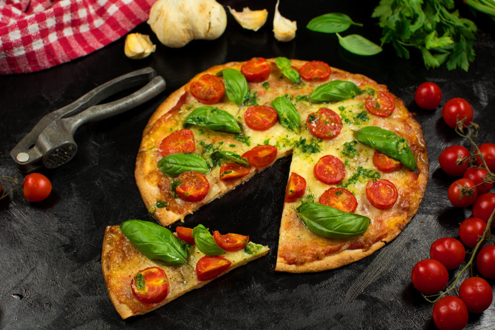 A freshly baked keto pizza topped with sliced cherry tomatoes and basil sits on a dark surface, with a slice removed and nearby garlic, cherry tomatoes, and a red-checkered cloth.