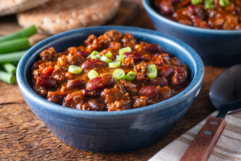 A blue bowl filled with hearty chili featuring ground meat and kidney beans, topped with chopped green onions, sits on a wooden table next to green onions and a spoon.