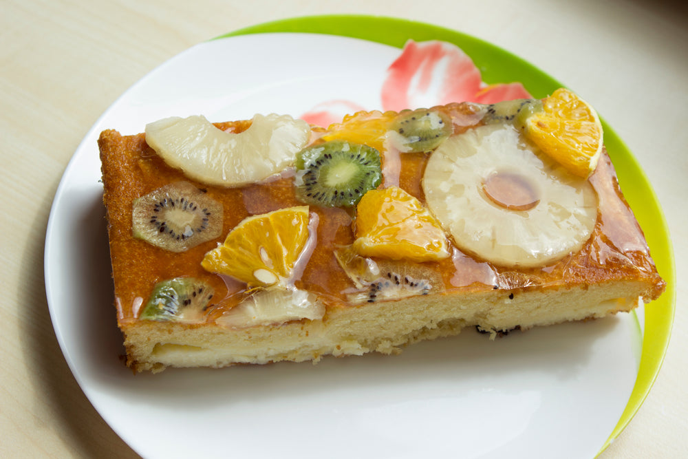 A slice of cake topped with assorted fruits, including pineapple, kiwi, and orange, is displayed on a white plate with a green rim, showcasing a glossy glaze.