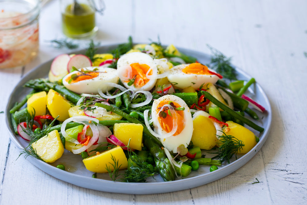 A colorful plate of egg salad features sliced hard-boiled eggs, asparagus, yellow potatoes, radishes, and herbs, garnished with red chili and served on a light wooden table.