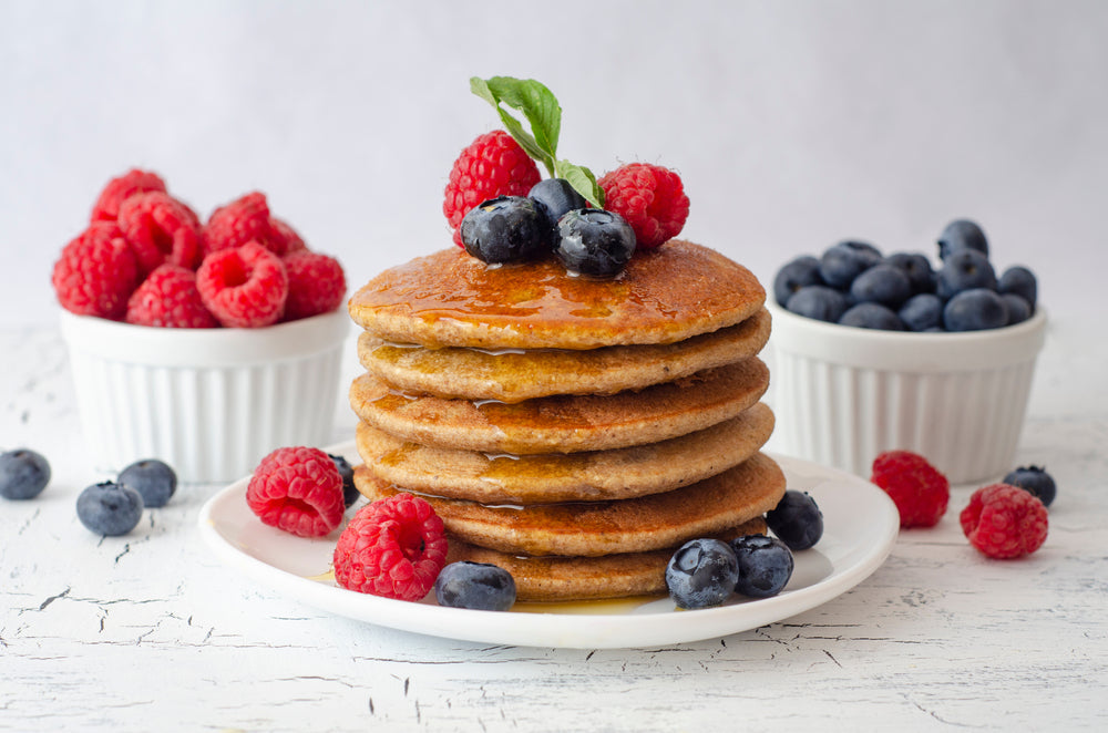 A stack of golden-brown oatmeal pancakes drizzled with syrup is topped with fresh blueberries and raspberries, surrounded by bowls of more berries on a light wooden surface.