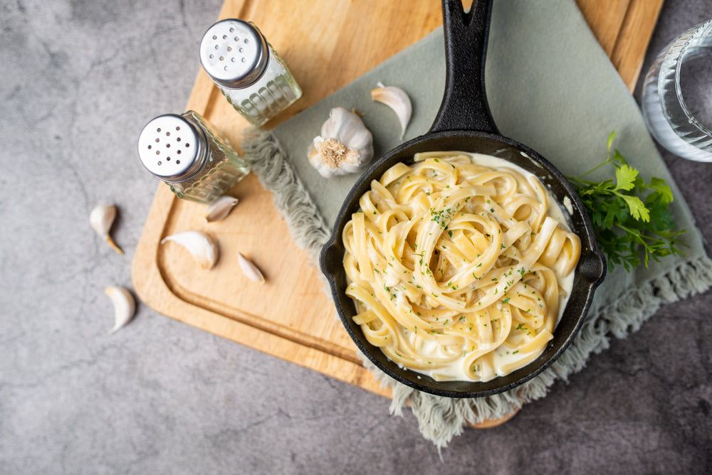 A cast iron skillet holds creamy fettuccine Alfredo topped with parsley, surrounded by garlic cloves, salt and pepper shakers, and a glass of water on a wooden cutting board.