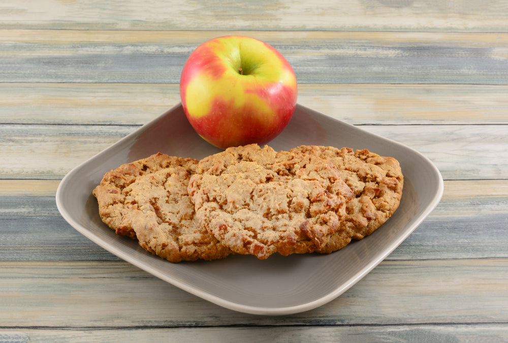 A gray plate holds two golden-brown apple cinnamon oatmeal cookies beside a red and green apple, set against a rustic wooden background.