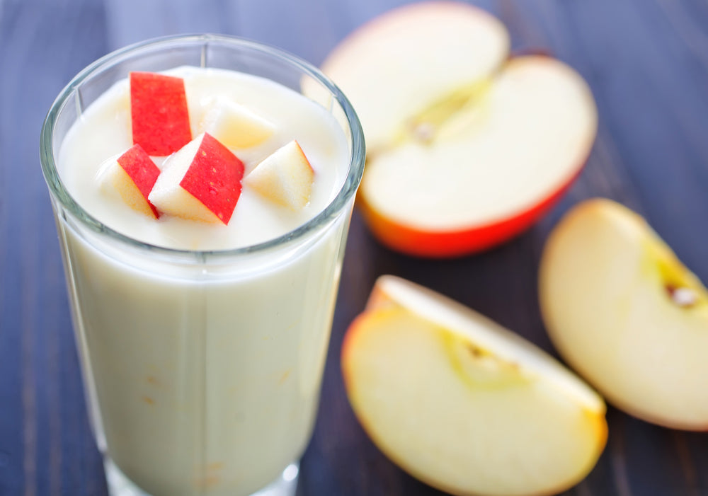 A tall glass of creamy apple pie shake topped with small red apple pieces sits on a wooden surface, accompanied by sliced red apples in the background.