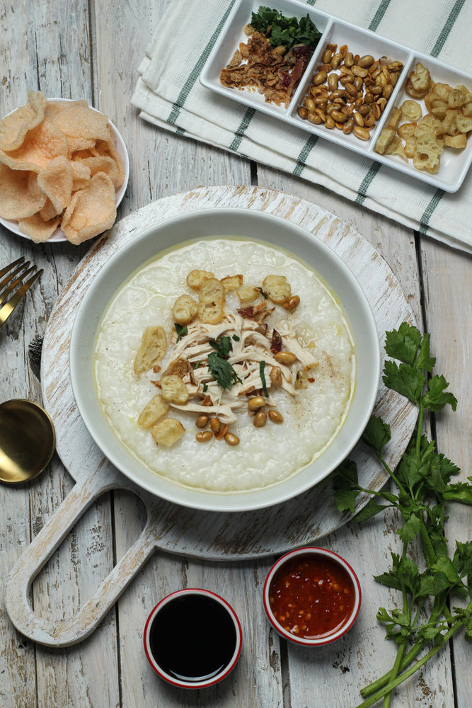A bowl of creamy mushroom soup topped with shredded chicken, crispy toppings, and herbs is surrounded by dipping sauces, snacks, and fresh cilantro on a rustic wooden table.