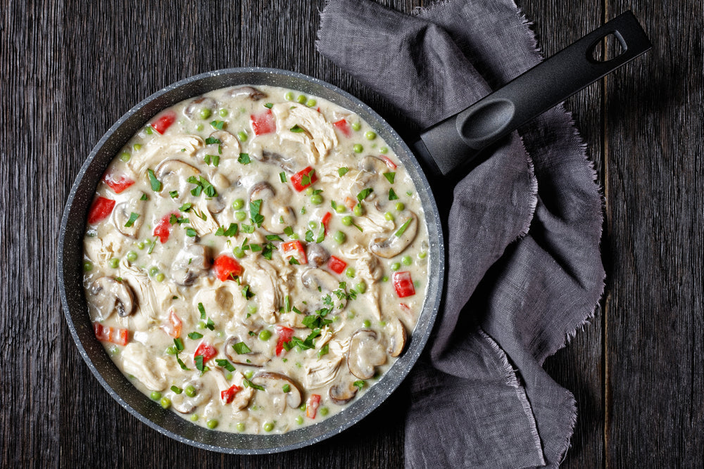 A black skillet filled with creamy A la King sauce, featuring chicken, mushrooms, red peppers, and green peas, sits on a wooden surface next to a folded gray cloth.
