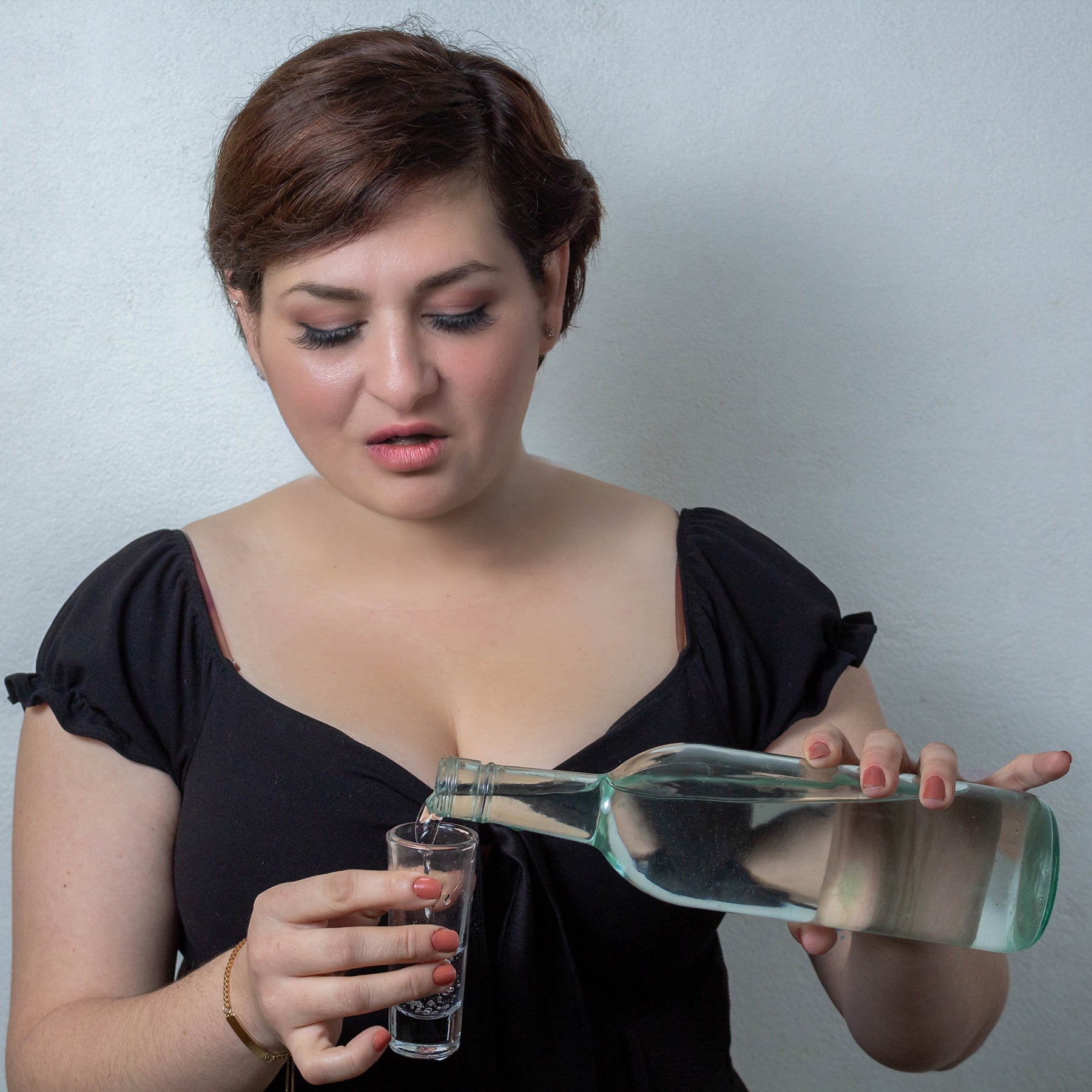 A woman with short hair is pouring water from a glass bottle into a shot glass while looking down thoughtfully, against a plain white background.