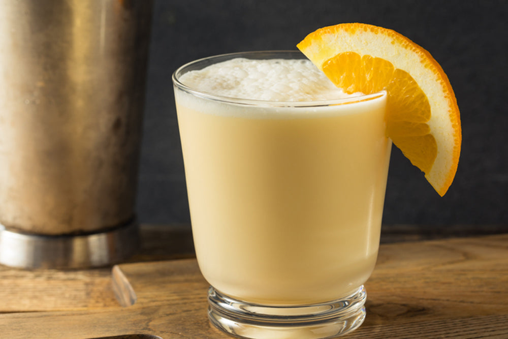 A frothy, pale orange drink is served in a clear glass, garnished with a slice of orange, beside a metal shaker on a wooden surface.
