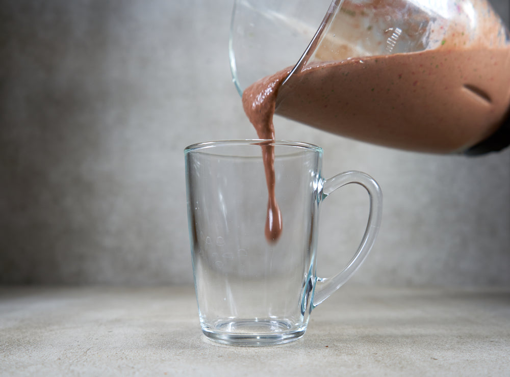 A glass measuring cup is pouring a thick, chocolate-colored smoothie into a clear glass mug on a light gray countertop.
