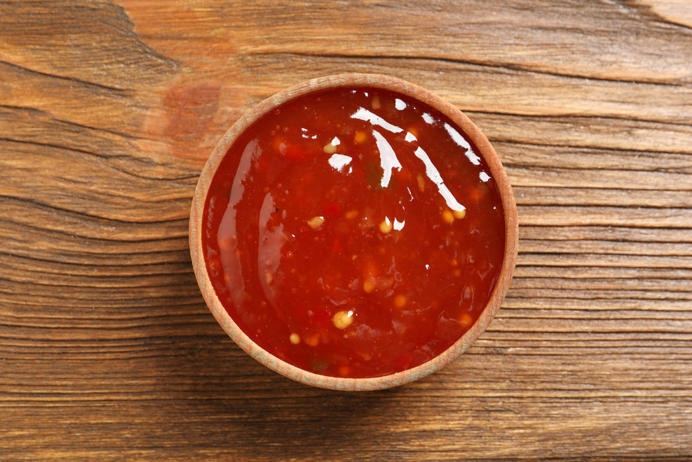 A round wooden bowl contains a glossy, red sweet and sour sauce, sitting on a textured wooden surface.