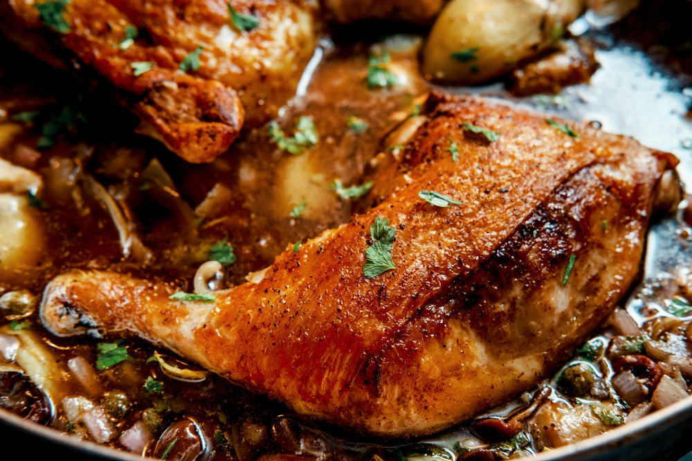 Golden-brown chicken pieces are simmering in a rich sauce with herbs and vegetables, garnished with fresh parsley, in a dark skillet.