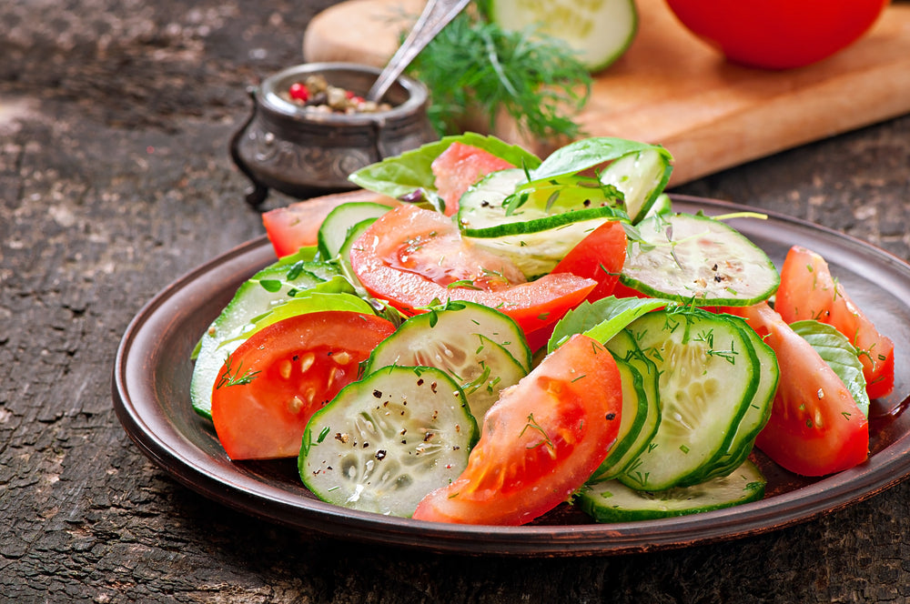 A colorful salad of sliced tomatoes and cucumbers garnished with fresh herbs is presented on a dark wooden table, with a bowl of spices in the background.