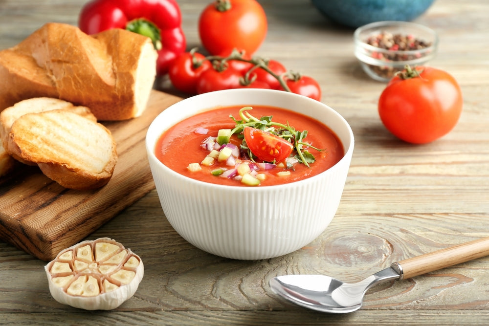 A bowl of vibrant red gazpacho is garnished with diced vegetables and herbs, accompanied by slices of bread and garlic on a wooden cutting board, with fresh tomatoes in the background.