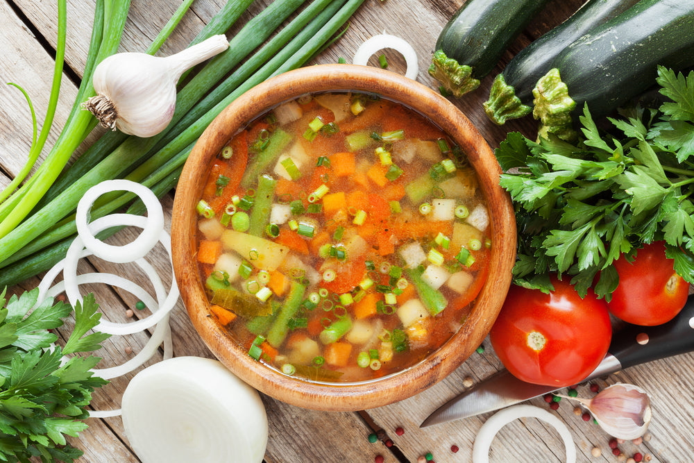 A wooden bowl filled with colorful zucchini soup sits on a rustic wooden surface, surrounded by fresh vegetables like garlic, onions, tomatoes, and herbs, ready for preparation.