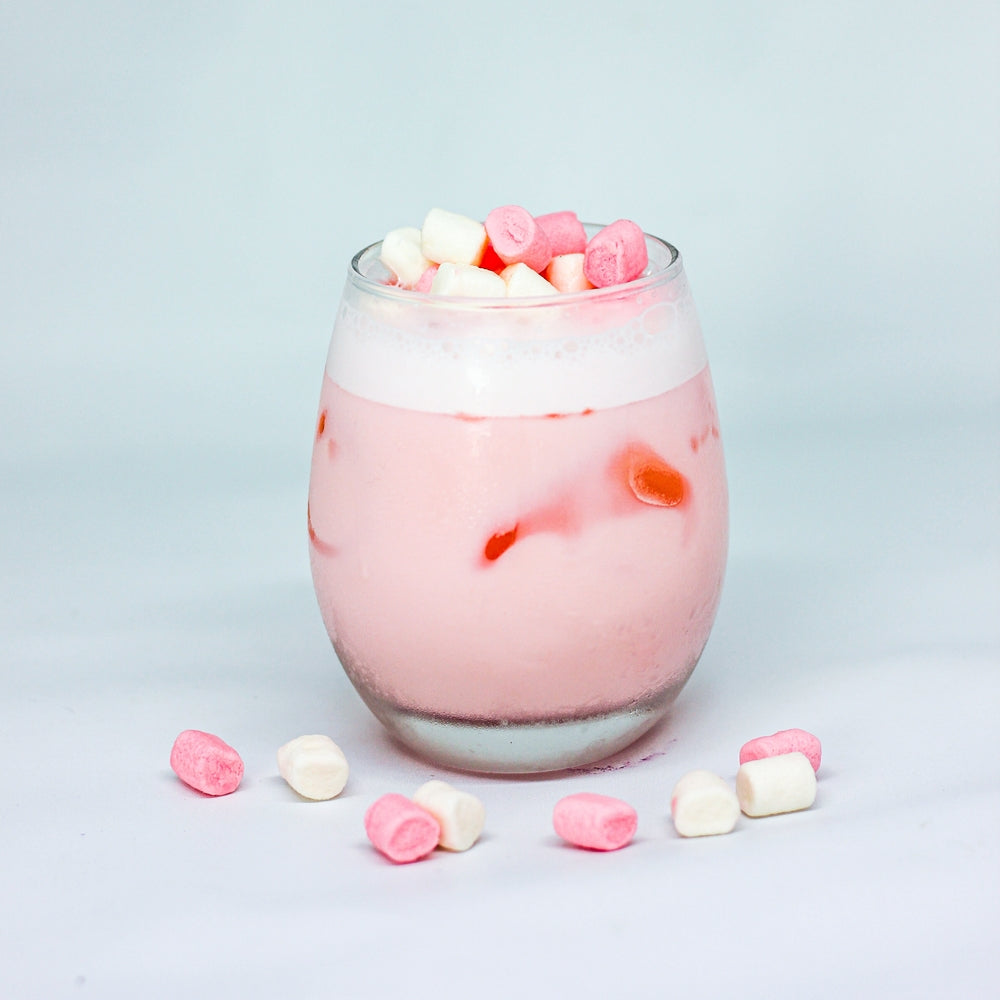 A glass filled with a pink, creamy shake topped with colorful mini marshmallows sits on a white surface, surrounded by scattered marshmallows in pink and white.