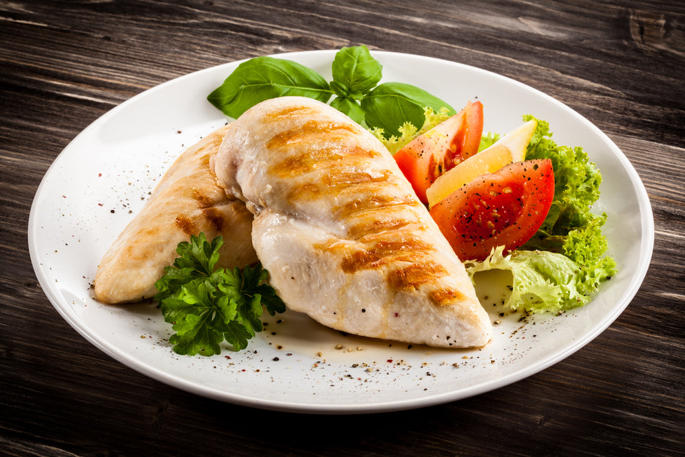 Grilled chicken breasts are served on a white plate with fresh basil, lettuce, sliced tomatoes, and lemon, set against a dark wooden background.