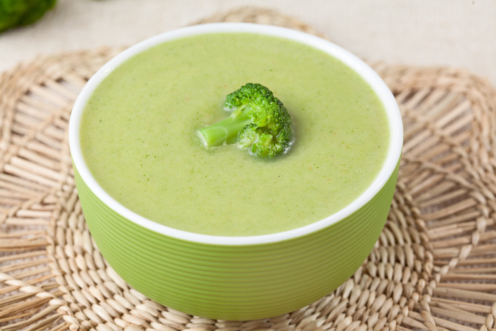 A green bowl filled with creamy broccoli soup sits on a woven placemat, topped with a small piece of broccoli, highlighting its fresh and vibrant color.