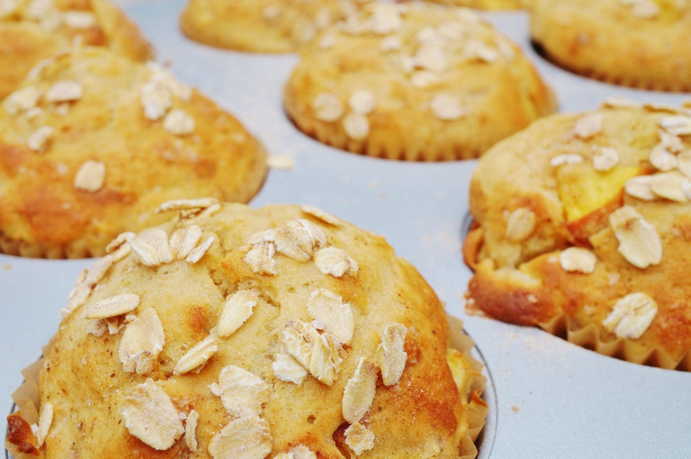 Golden-brown peach cobbler muffins topped with oats are arranged in a muffin tin, showcasing their fluffy texture and inviting appearance.