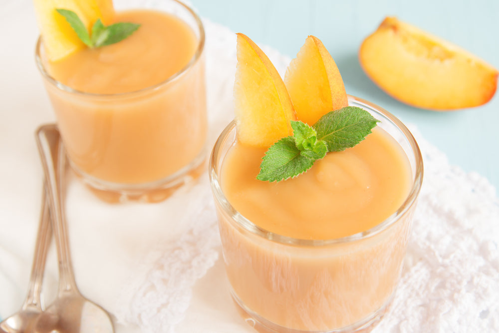 Two glasses of peach smoothie, topped with peach slices and mint leaves, sit on a white lace napkin with silver spoons, against a light blue background.