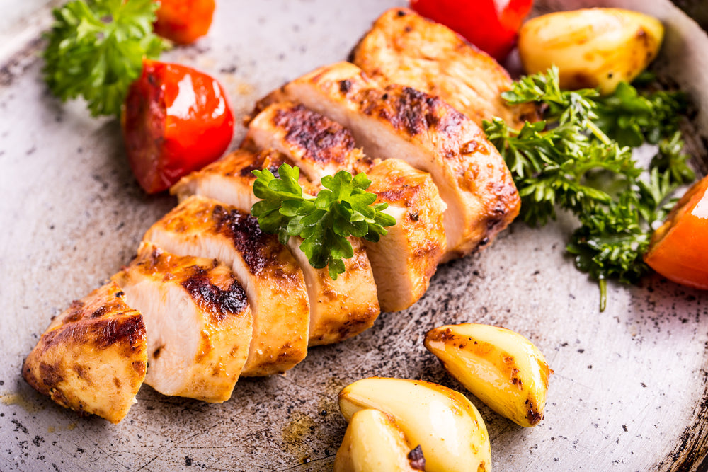 Sliced, golden-brown baked chicken is garnished with fresh parsley, accompanied by roasted garlic and cherry tomatoes on a rustic plate.