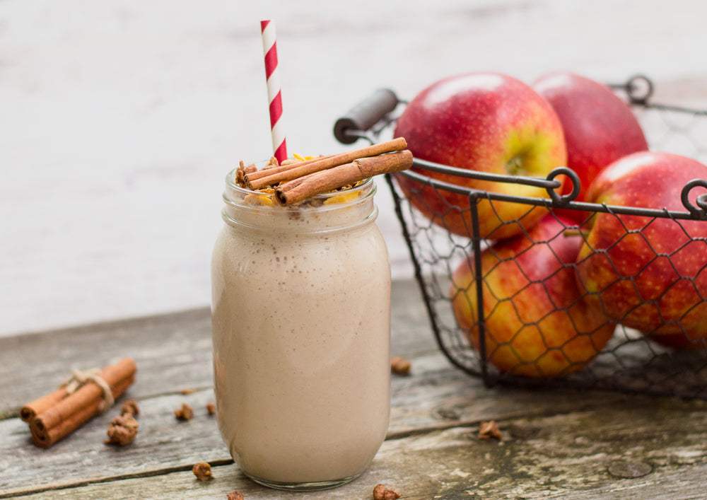 A glass jar filled with a creamy shake topped with cinnamon sticks and colorful sprinkles sits on a wooden table, with a wire basket of red apples in the background.