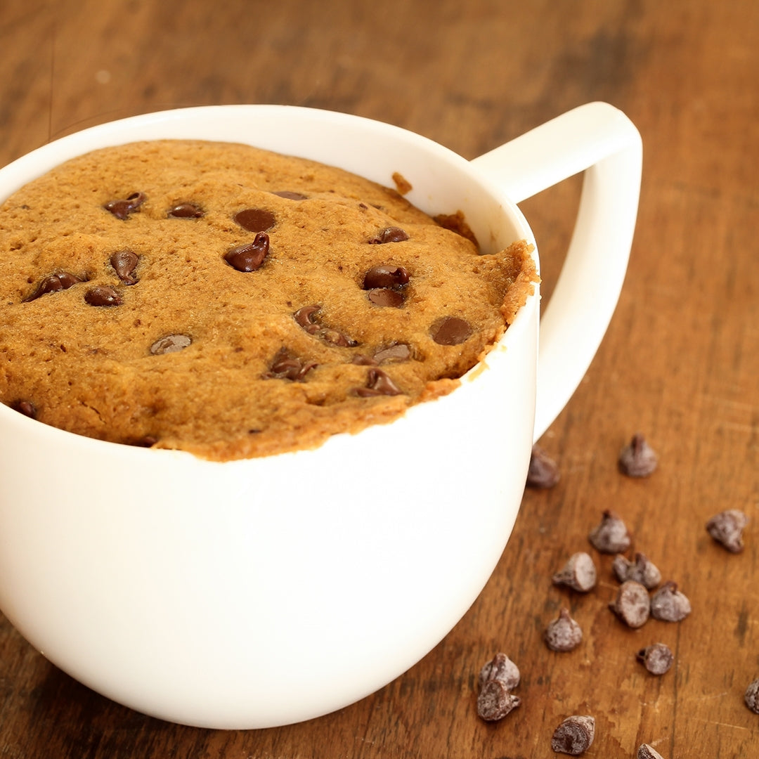 A white mug filled with a warm, gooey chocolate chip cookie sits on a wooden surface, with scattered chocolate chips nearby, representing a dessert option after bariatric surgery.