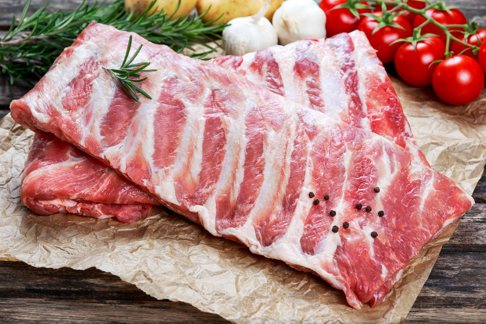 Raw beef ribs are arranged on crumpled parchment paper, garnished with a sprig of rosemary and black pepper, with fresh tomatoes, garlic, and potatoes in the background on a wooden surface.