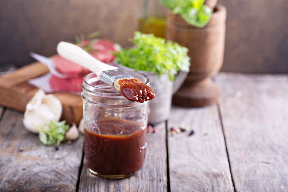 A jar of barbecue sauce sits on a wooden table, with a brush dipped in the sauce resting on the rim, surrounded by fresh herbs, garlic, and raw meat in a rustic kitchen setting.