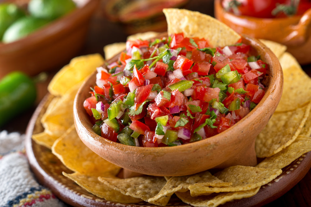 A clay bowl filled with vibrant salsa made of diced tomatoes, onions, and green peppers is surrounded by crispy tortilla chips on a wooden platter, with fresh limes in the background.