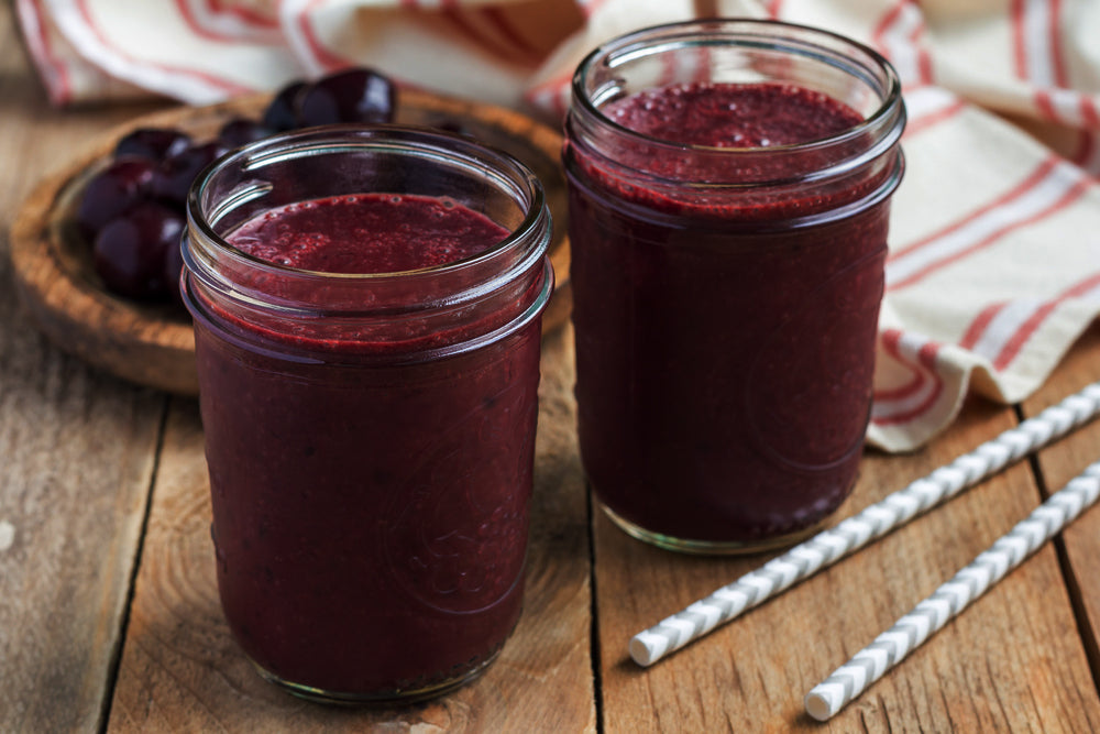Two jars filled with dark purple cherry smoothie sit on a wooden table, accompanied by a bowl of cherries and striped straws, suggesting a refreshing drink option.