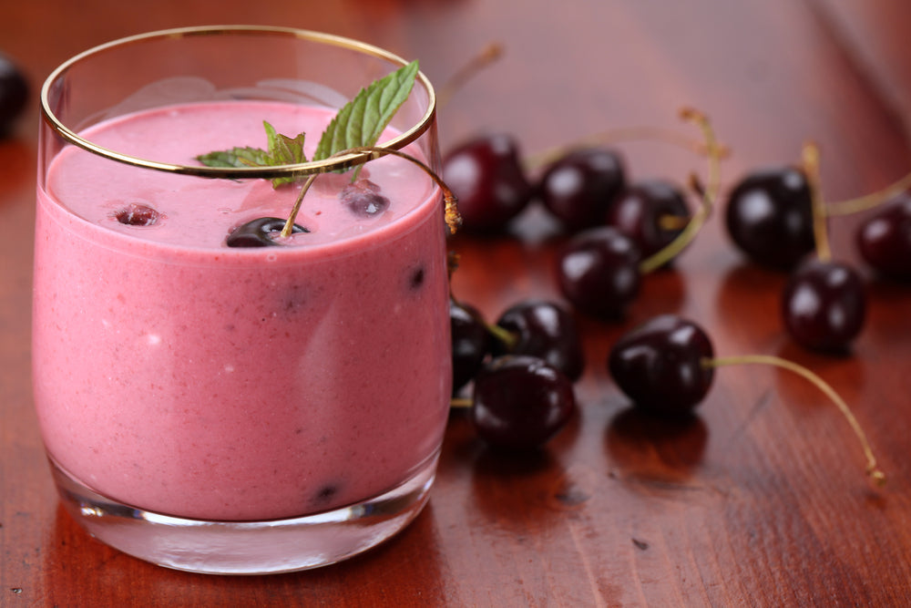 A glass of pink cherry smoothie garnished with a mint leaf sits on a wooden surface, surrounded by fresh cherries, showcasing a refreshing drink.
