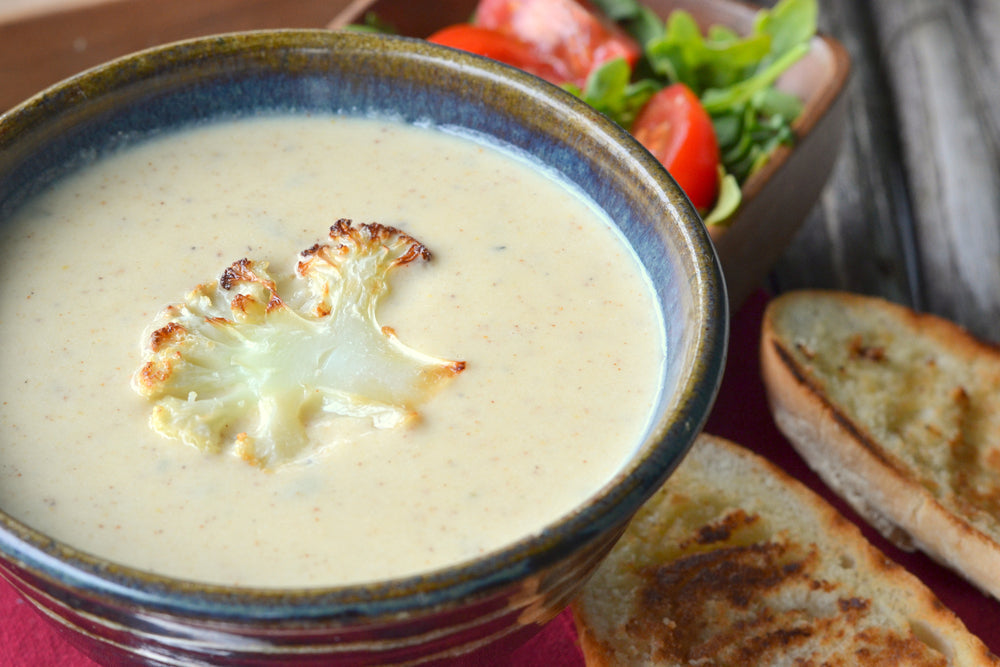 A bowl of creamy cauliflower soup topped with a roasted cauliflower floret sits alongside slices of toasted bread and a small salad with tomatoes and greens on a wooden table.