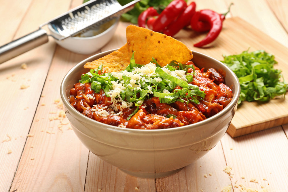 A bowl of turkey chili topped with shredded cheese and fresh cilantro, garnished with a tortilla chip, sits on a wooden table alongside fresh herbs and red chili peppers.