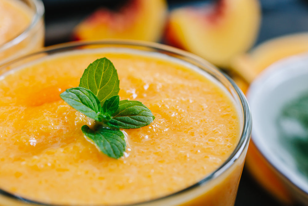 A close-up of a vibrant orange smoothie topped with a fresh mint leaf, surrounded by sliced peaches, set against a dark background.