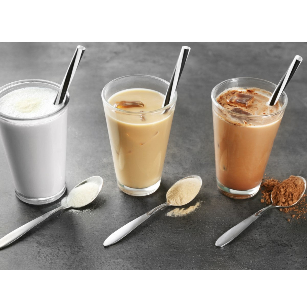 Three glasses of meal replacement shakes in varying colors—white, beige, and brown—are placed on a dark surface, accompanied by silver spoons with powder beside each glass.