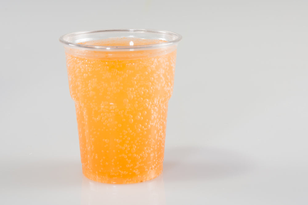 A clear plastic cup filled with a bubbly, bright orange drink sits on a light surface, suggesting a refreshing peach mango cooler beverage.