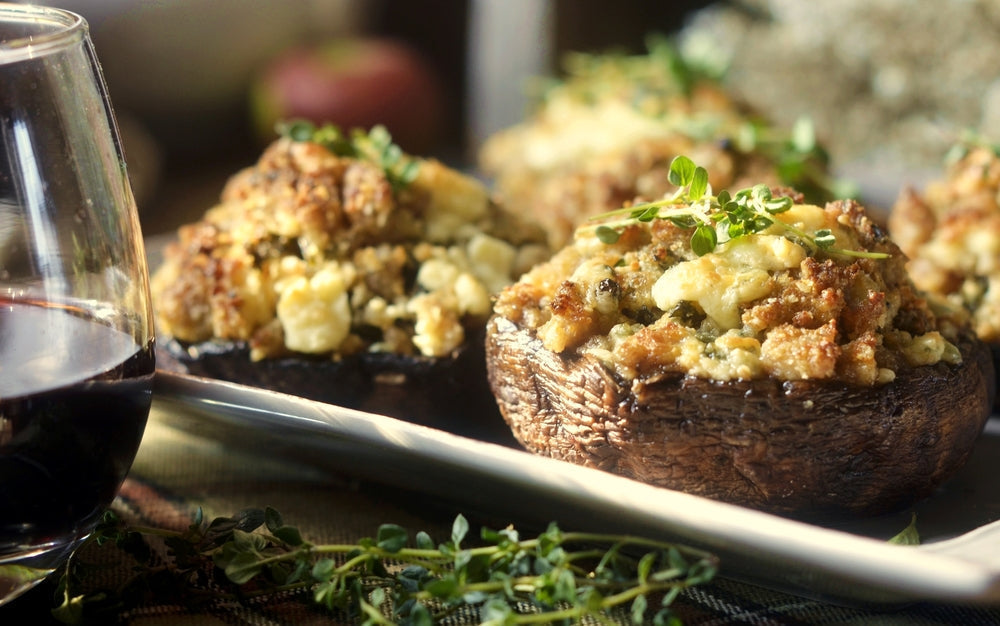 Three stuffed mushrooms topped with herbs sit on a platter next to a glass of red wine, with a blurred background suggesting a cozy dining setting.