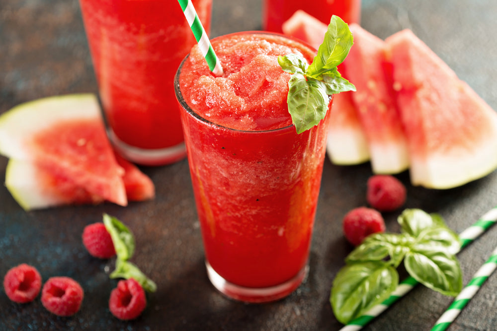 A vibrant raspberry slush in a tall glass, topped with a fresh basil leaf and a striped straw, is surrounded by watermelon slices and fresh raspberries on a dark surface.