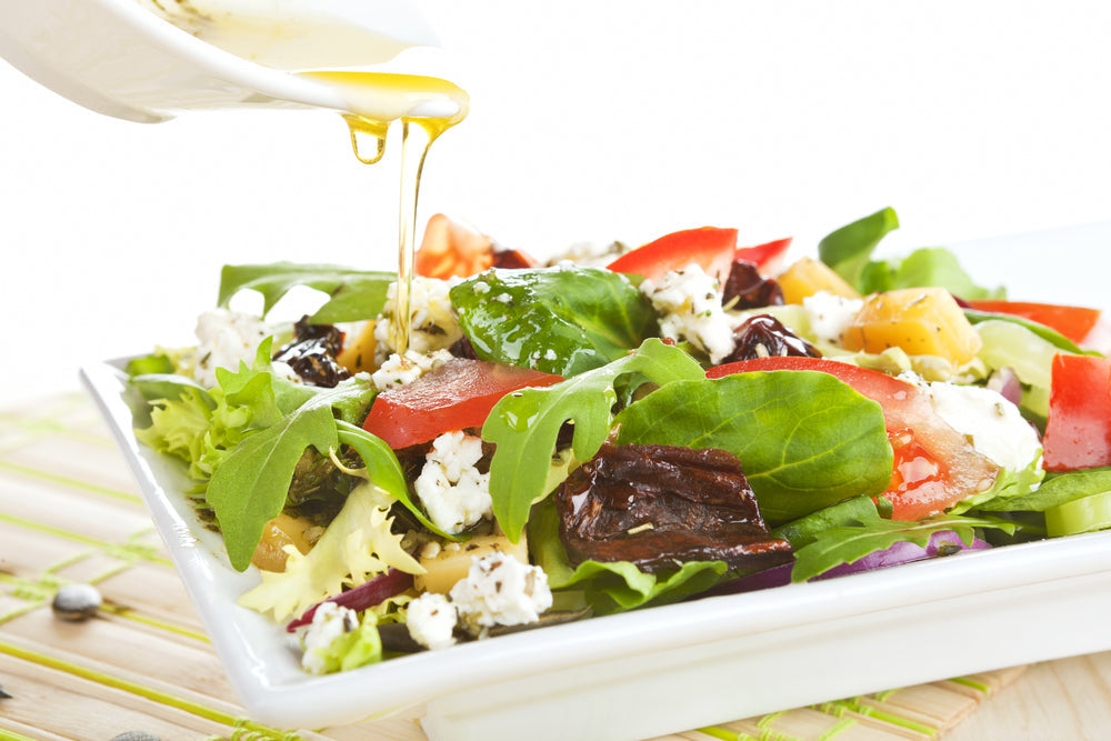A creamy salad dressing is being poured over a colorful salad filled with mixed greens, tomatoes, cheese, and dried fruits, presented on a white plate against a light background.