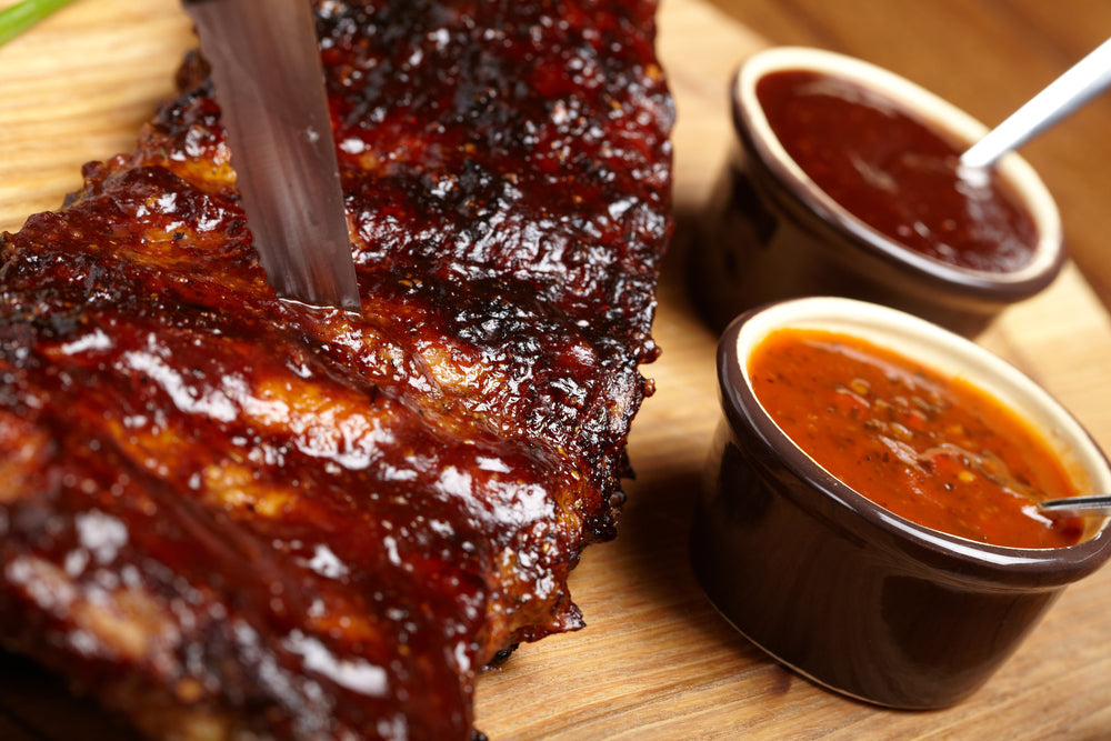 A close-up of a juicy, glazed barbecue rib being sliced with a knife, accompanied by two small bowls of different barbecue sauces on a wooden cutting board.