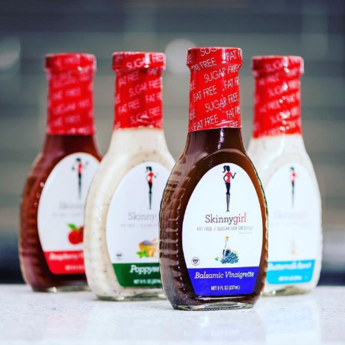 Four bottles of Skinnygirl salad dressings are displayed on a countertop, featuring flavors like Balsamic Vinaigrette and Poppyseed, each with a bright red cap labeled "Fat Free" and "Sugar Free."