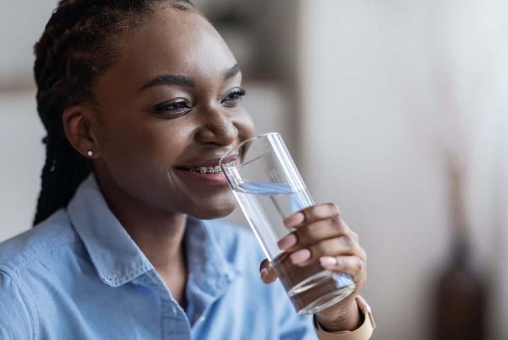 A smiling woman with braces holds a glass of water close to her mouth, sitting in a bright room that suggests a focus on hydration after bariatric surgery.