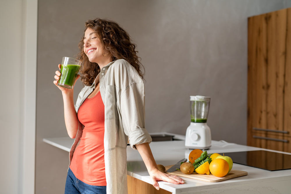 A woman with curly hair smiles while holding a glass of green smoothie in a modern kitchen, with a blender and fresh fruits on the counter, emphasizing healthy hydration after weight loss surgery.