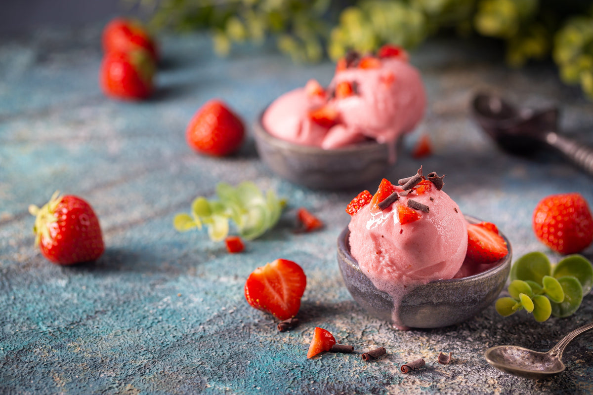 Two bowls of pink strawberry soft serve ice cream are topped with fresh strawberries and chocolate shavings, surrounded by whole strawberries and green leaves on a textured blue surface.