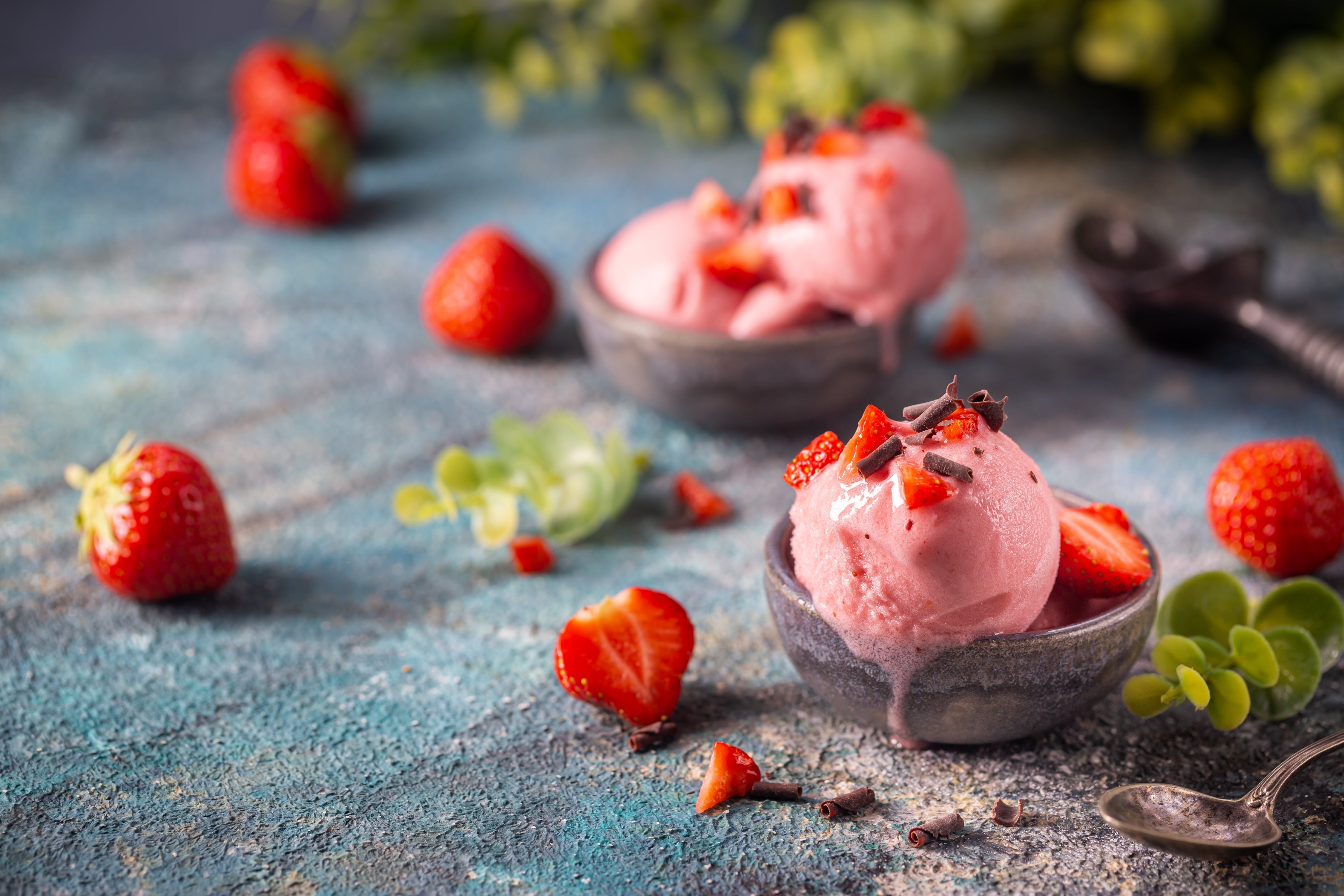 Two bowls of pink strawberry soft serve ice cream are topped with fresh strawberries and chocolate shavings, surrounded by whole strawberries and green leaves on a textured blue surface.