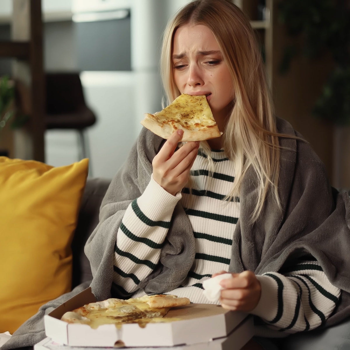 A young woman wrapped in a gray blanket looks distressed while holding a slice of pizza, sitting on a couch with a pizza box and a yellow pillow nearby, reflecting a moment of stress eating.